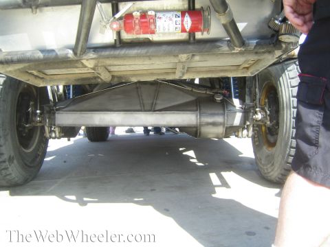 1990 Ford Bronco - Low-angle underside view of an off-road vehicle revealing its rear axle, exhaust muffler, and a mounted red fire-extinguisher bottle.