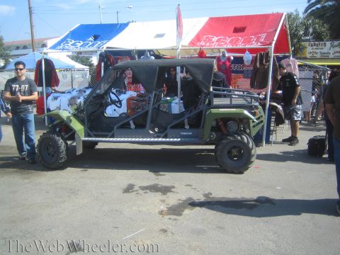 1990 Ford Bronco - green four-seat tube-frame off-road buggy sits among red and blue vendor tents, with a man standing at left.