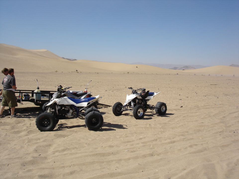 Two white and blue Suzuki sport quads with paddle tires parked on sand dunes near a trailer