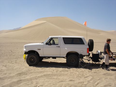 1996 Ford Bronco - A white 1996 Ford Bronco is parked on a sandy dune under a clear blue sky, with a person standing nearby.