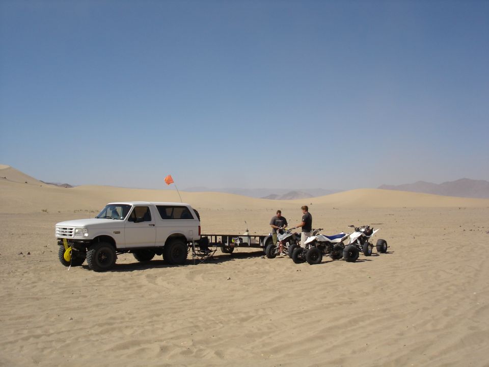 White lifted Ford Bronco with trailer and three quads parked in desert sand dunes