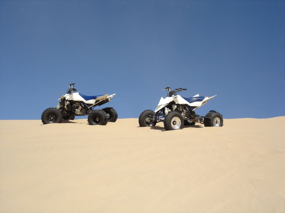 Two white and blue Suzuki sport quads with paddle tires on a dune crest against blue sky