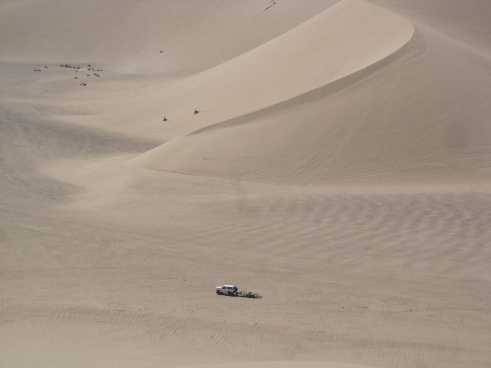 White SUV and trailer parked in a huge sand bowl with quads riding the dune ridge above