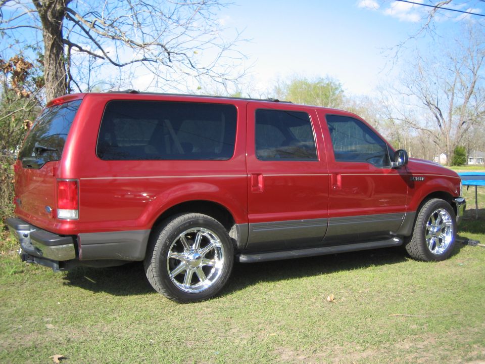 Red Ford Excursion SUV with chrome wheels parked on grass, rear passenger-side view