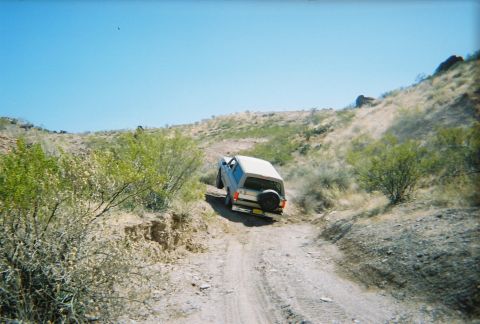 1996 Ford Bronco - A beige SUV navigates a rough, unpaved path through a dry, arid landscape under a clear blue sky.