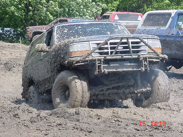 Mud-covered Ford Bronco with tube bumper plowing through deep mud at a mud pit, dated 15