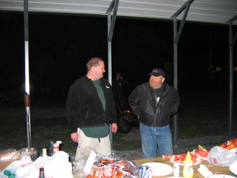 1985 Ford LTD Crown Victoria - Two men converse near a picnic table laden with food, under a covered structure at night.