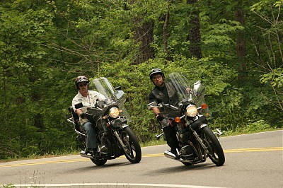 1985 Ford LTD Crown Victoria - Two riders on motorcycles navigate a curve on a tree-lined road.