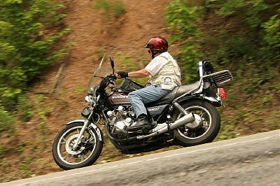 1985 Ford LTD Crown Victoria - A rider on a motorcycle navigates a winding road with a wooded hillside in the background.
