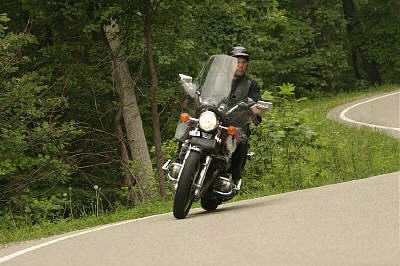 1985 Ford LTD Crown Victoria - A rider on a motorcycle navigates a curve on a tree-lined road.
