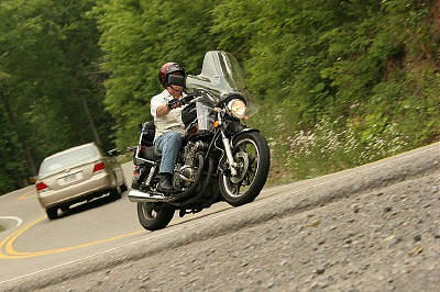 1985 Ford LTD Crown Victoria - A rider leans into a curve on a motorcycle, with a car following on a tree-lined road.