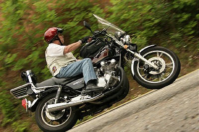 1985 Ford LTD Crown Victoria - A rider on a motorcycle navigates a curved road surrounded by trees.