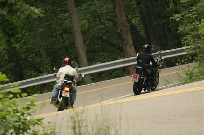 1985 Ford LTD Crown Victoria - Two motorcyclists ride on a scenic road surrounded by trees.