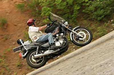 1985 Ford LTD Crown Victoria - A rider on a motorcycle navigates a paved road surrounded by foliage.