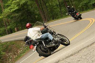 1985 Ford LTD Crown Victoria - Two motorcycles navigate a sharp curve on a tree-lined road, with one rider leaning low.