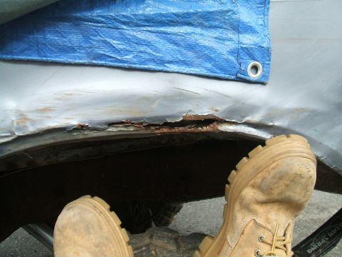 1995 Ford Bronco - The wheel well of a vehicle shows significant rust and damage, partially covered by a blue tarp. Tan work boots are visible in the foregrou…