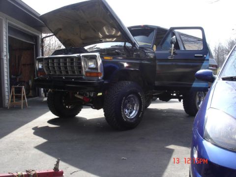 1979 Ford Bronco - A black Ford Bronco is parked outside a garage with its hood and driver's door open. A blue car is partially visible on the right.