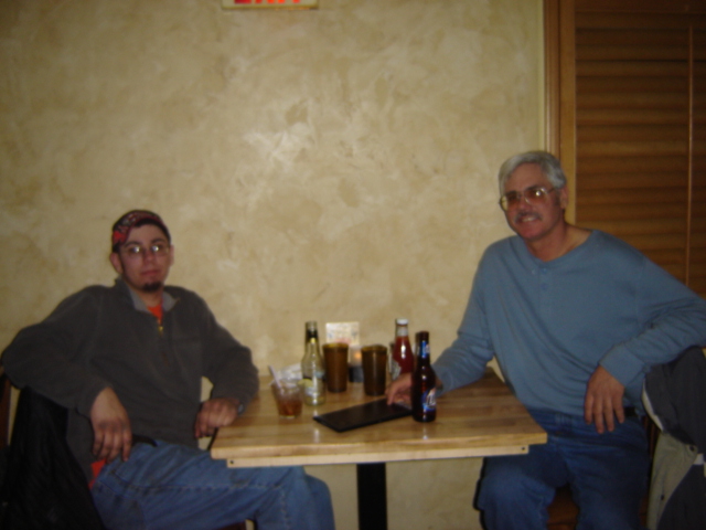 Two men sit at a wooden table with drinks and bottles, with a textured wall behind them