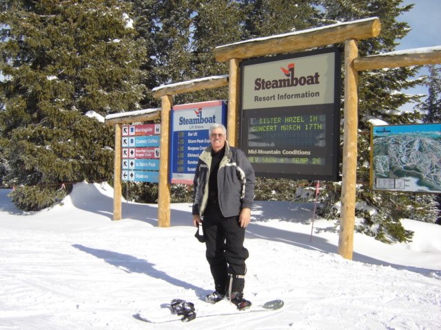 Man in ski gear stands on a snowboard in front of Steamboat Resort signs