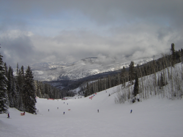 Ski slope with scattered skiers, snow-covered trees, and misty mountains under a cloudy sky. Watermark reads SUPERMOTORS
