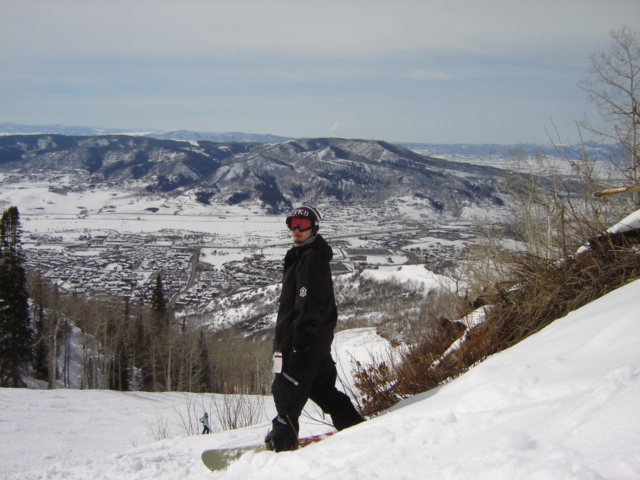 Snowboarder on snowy slope with town and mountains in background. Supermotors.net logo visible