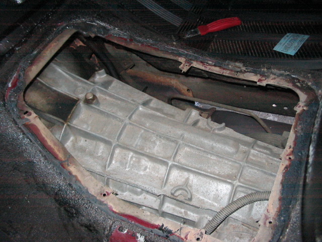 Interior view of a vehicle's transmission tunnel with the cover removed, showing the transmission casing. A red-handled tool is visible on the dashboard