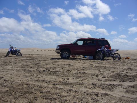 1996 Ford Explorer - A red SUV and two dirt bikes are parked on a sandy landscape under a cloudy sky.