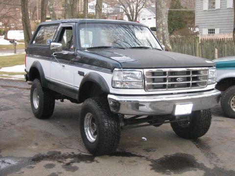 1994 Ford Bronco - A two-tone lifted Ford Bronco is parked on a paved surface with another vehicle partially visible to the right.
