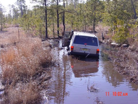 1995 Dodge 1500 - A white Jeep Cherokee navigates a muddy, water-filled trail with its driver's door ajar.