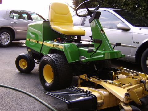 1996 Mercury Grand Marquis - A green John Deere F710 lawn tractor is parked on asphalt next to a silver sedan.