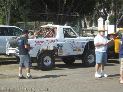 1990 Ford Bronco - white Ford Bronco off-road race truck lettered "Anger Issues" with number 349 parked under trees while two men inspect it.