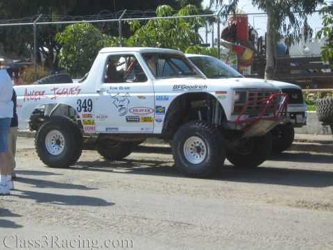 1990 Ford Bronco - white "Anger Issues" number 349 Ford Bronco off-road race truck sits on a dirt lot with a man standing at left.