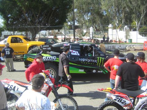 1990 Ford Bronco - black and green Monster Energy KMC Wheels number 27 off-road truck is staged among spectators, with dirt bikes in the foreground.
