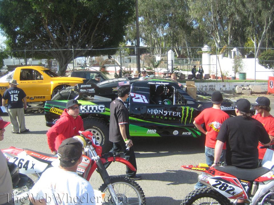 Black and green Monster Energy number 27 off-road truck among spectators and bikes