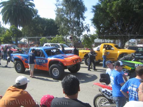 1990 Ford Bronco - orange-and-blue Fabtech truck and a yellow Greer Bros truck staged in line as a crowd of spectators watches.