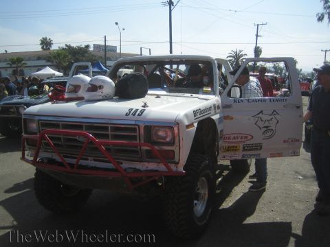 1990 Ford Bronco - Side-front view of white race truck 349 with two helmets resting on the hood, a red tube bumper and BFGoodrich decals, surrounded by specta…
