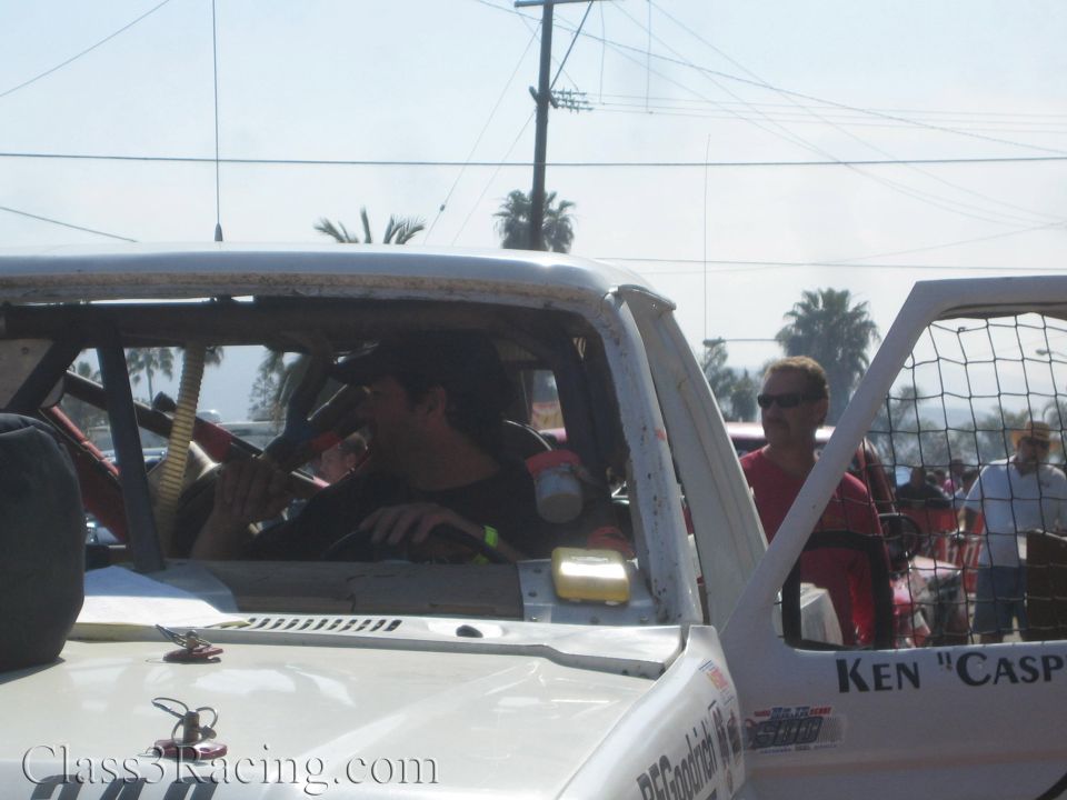 Driver seated inside the cab of a white off-road race truck wiping his face