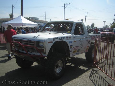 1990 Ford Bronco - lifted white Ford off-road race truck numbered 349 sits on large knobby tires behind a red barrier amid a crowded outdoor event.