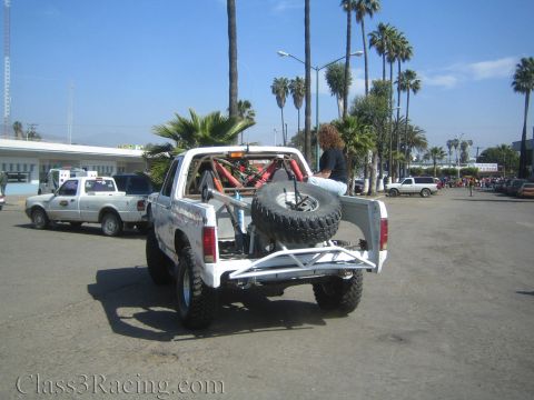 1990 Ford Bronco - Rear three-quarter view of a white off-road race truck with a spare tire in the bed and tube cage, parked on pavement lined with palm trees.