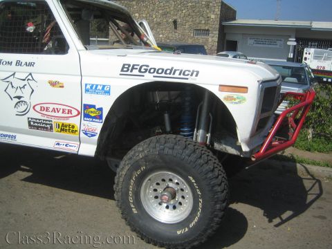 1990 Ford Bronco - Close view of a white off-road truck's front corner showing a BFGoodrich tire, blue coilover shock, red tube bumper and sponsor decals.