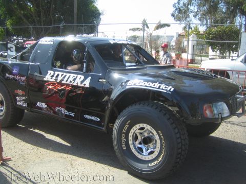 1990 Ford Bronco - black off-road race truck lettered 'Riviera Racing' with a smooth fiberglass body sits on BFGoodrich Baja tires as spectators stand nearby.