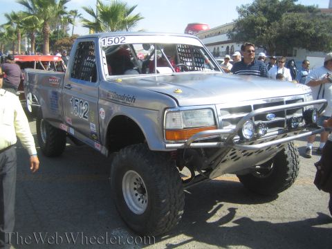 1990 Ford Bronco - silver Ford off-road race truck numbered 1502 with chrome brush guard, roof light bar and large tires sits at a staging area near palm tree…