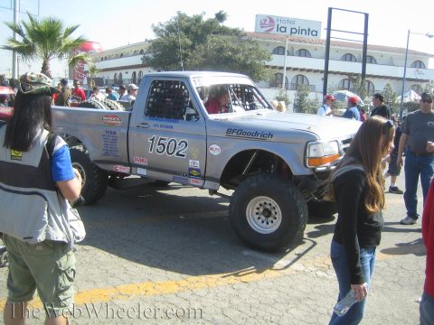 1990 Ford Bronco - Side view of silver Ford off-road race truck 1502 parked on a street in front of Hotel La Pinta as spectators walk past.