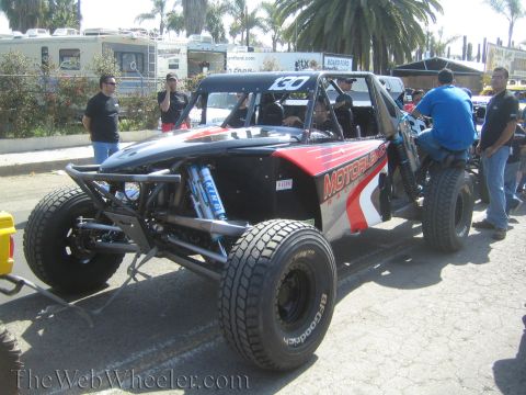 1990 Ford Bronco - black and red tube-chassis off-road race buggy sits on BFGoodrich tires with exposed long-travel suspension as crew work around it.