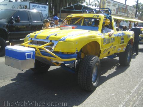 1990 Ford Bronco - yellow and blue off-road race truck numbered 719 with a cooler strapped to its front tube bumper sits near a Board Ford banner.