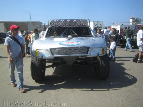 1990 Ford Bronco - Head-on view of a silver off-road race truck with a six-light roof bar and tube bumper, with people gathered around it on pavement.