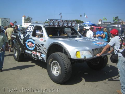 1990 Ford Bronco - silver off-road race truck numbered 55 with black tribal graphics and a roof-mounted light bar sits at staging as spectators look on.