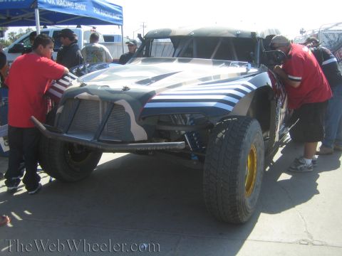 1990 Ford Bronco - Front view of a camouflage-painted off-road race truck with a tube bumper as two crew members in red work on either side under a canopy.