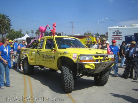 1990 Ford Bronco - yellow off-road race truck numbered 141 lettered 'Guzman Construction' with two pink lawn flamingos mounted on its roof in a parade lane.