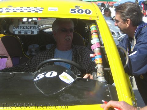 1990 Ford Bronco - gray-haired driver in a plaid shirt sits in yellow race buggy 500 talking with a man leaning at the open door.